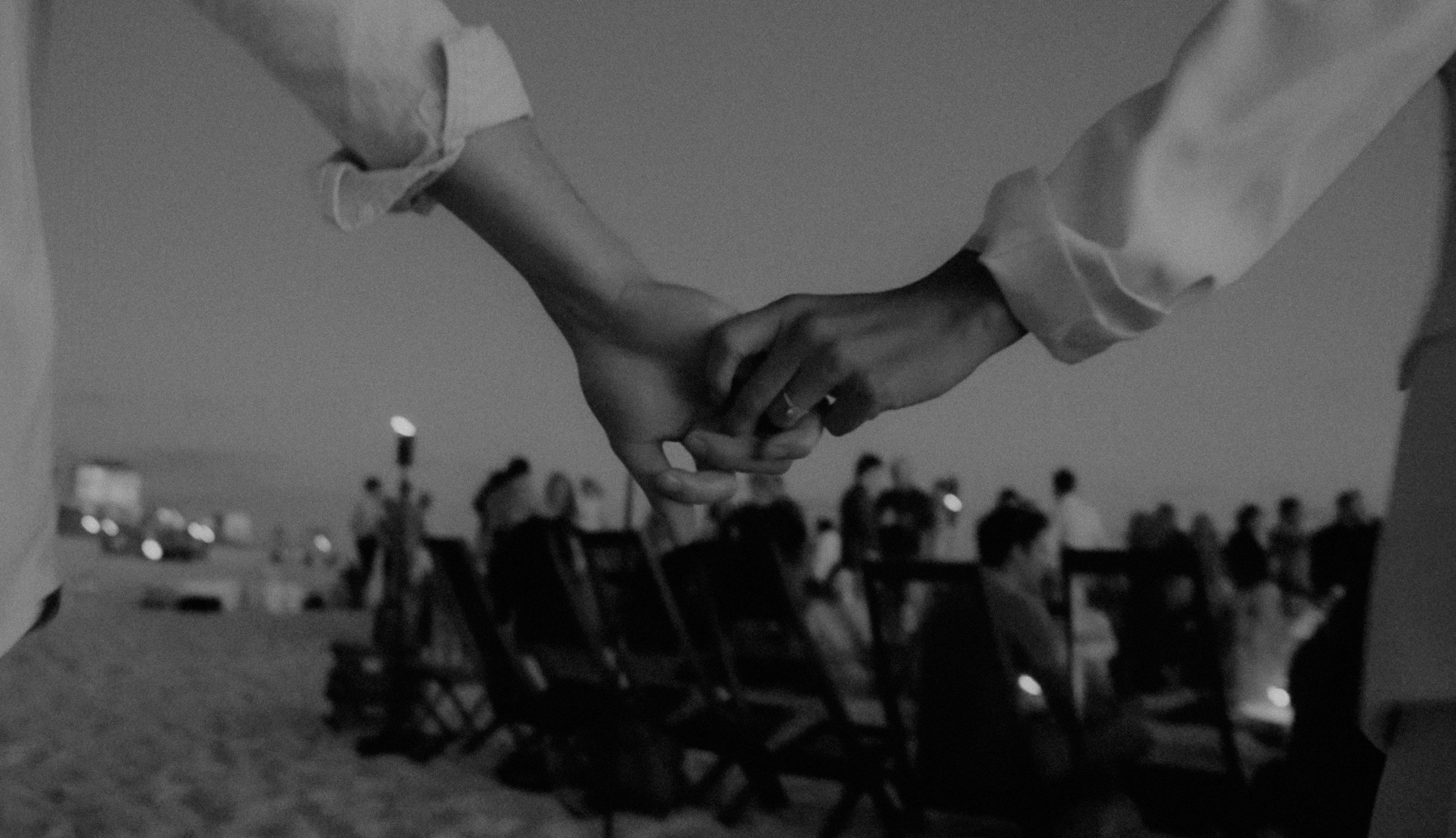 Black and white image of two people holding hands in the foreground and people sitting on beach chairs in the background. Image by - 550Park Luxury Wedding Films